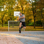 Man jumping in mid-air while wearing Fit Boots Rebound Boots on an outdoor basketball court surrounded by trees with autumn foliage.