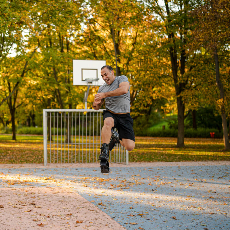 Man jumping in mid-air while wearing Fit Boots Rebound Boots on an outdoor basketball court surrounded by trees with autumn foliage.